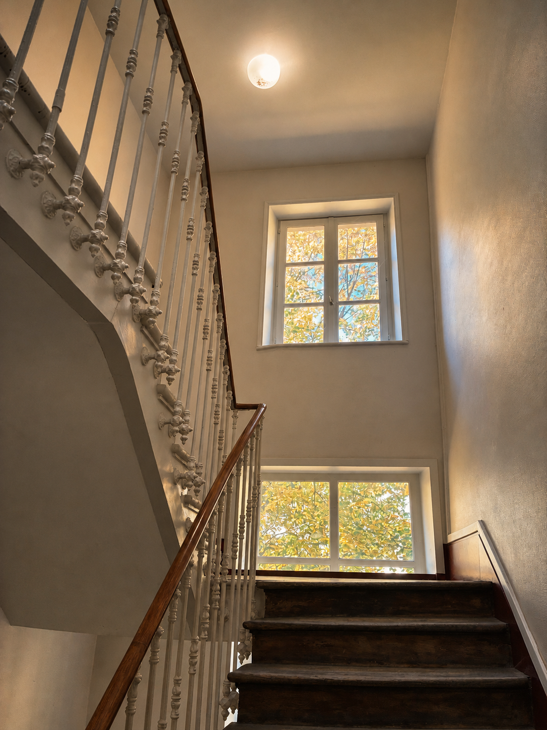 Cage d’escalier du lieu : escalier de bois, rampe claire ouvragée et fenêtres sur la verdure